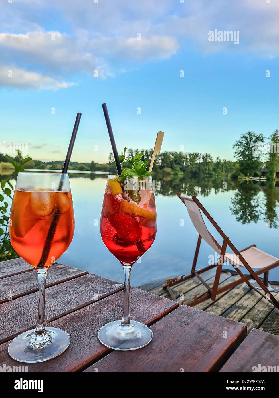 Due bicchieri di Aperol Spritz con fetta di paglia e limone su un tavolo, molo di legno vicino al lago con sdraio e vista sull'acqua, cielo blu con clou Foto Stock