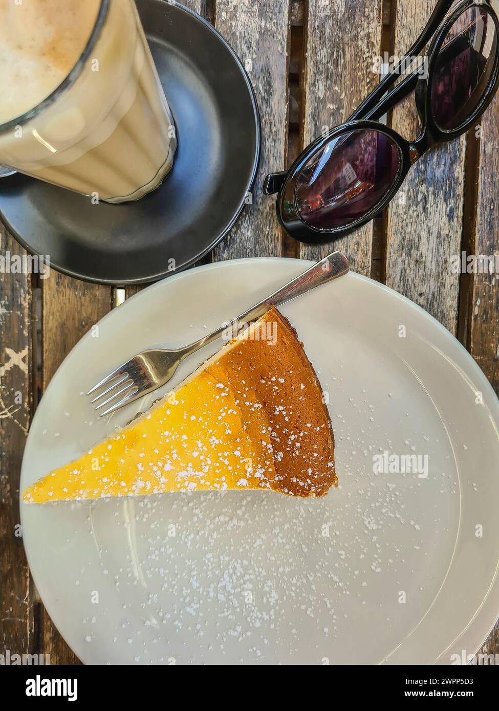 Un pezzo di cheesecake con zucchero a velo su un piatto di caffè bianco con latte in un bicchiere per dessert, occhiali da sole neri su un tavolo di legno Foto Stock