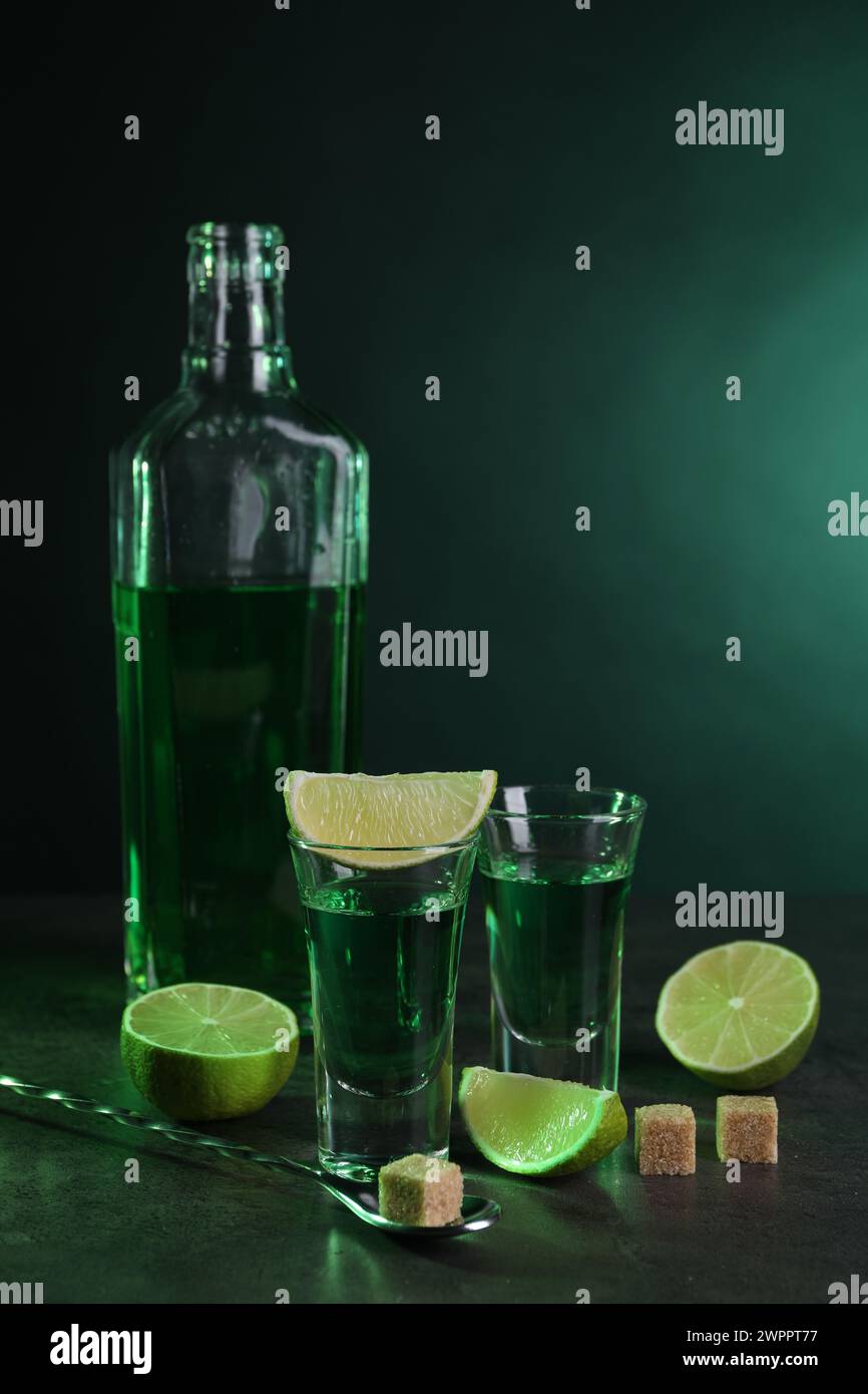 Absinthe in bicchieri da shot, cucchiaio, cubetti di zucchero di canna e lime su un tavolo testurizzato grigio. Bevanda alcolica Foto Stock