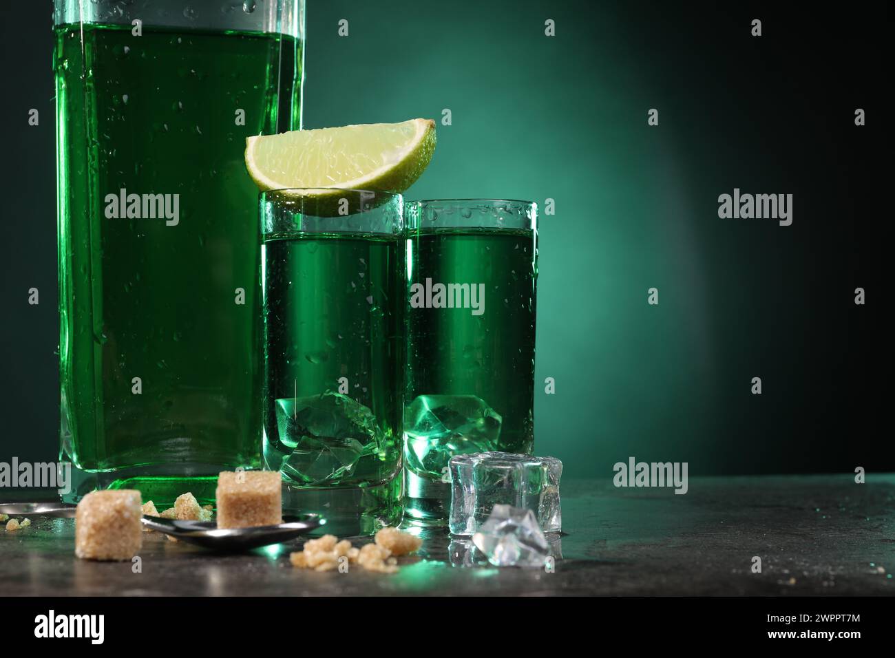 Absinthe in bicchieri da shot con cubetti di ghiaccio, lime, zucchero di canna e cucchiaio sul tavolo grigio su sfondo verde, spazio per il testo. Bevanda alcolica Foto Stock
