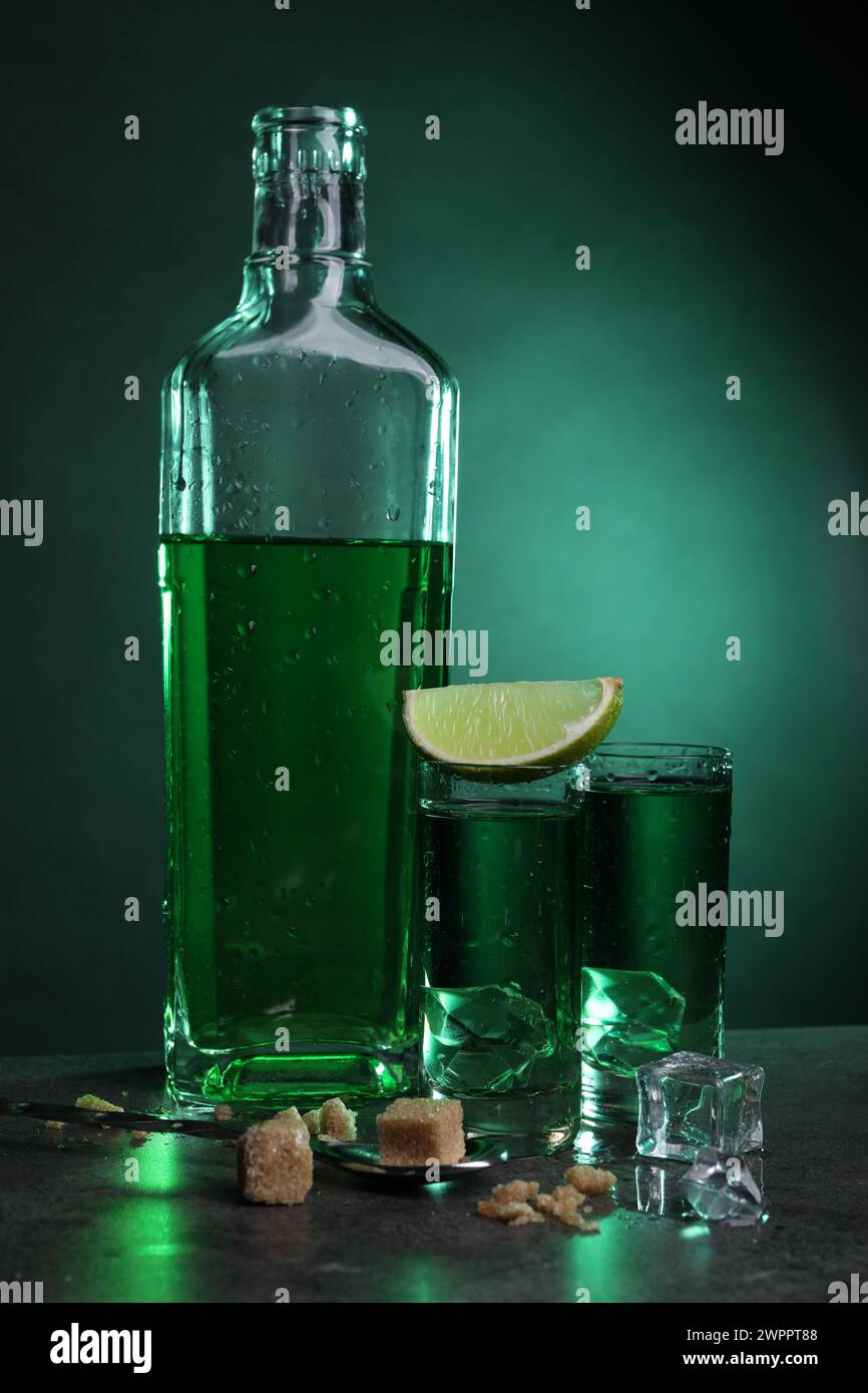 Absinthe in bicchieri da shot con cubetti di ghiaccio, lime, zucchero di canna e cucchiaio sul tavolo grigio su sfondo verde. Bevanda alcolica Foto Stock