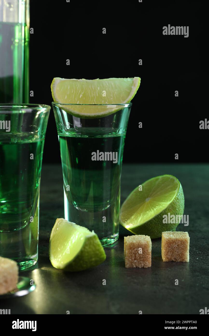 Absinthe in bicchieri da shot, lime e zucchero di canna a cubetti su tavolo grigio su sfondo nero. Bevanda alcolica Foto Stock