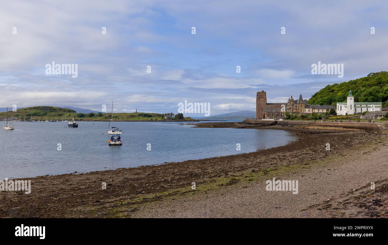 Barche ormeggiate punteggiano la baia di Oban accanto a una riva ricoperta di alghe, con la cattedrale di Santa Colomba e gli edifici del lungomare in lontananza, Scozia. Foto Stock
