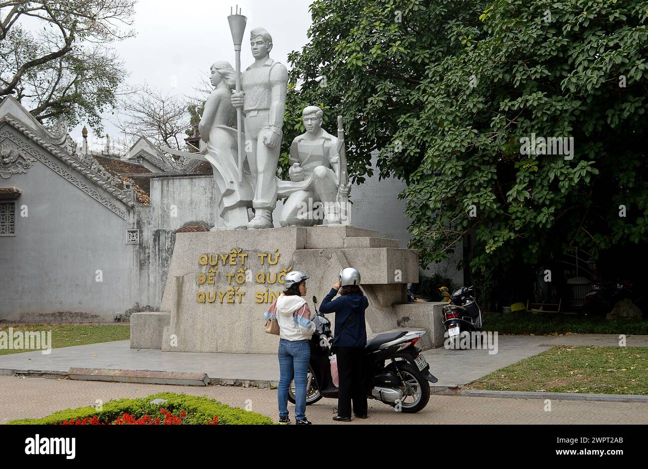 HANOI, VIETNAM - 14 febbraio 2024: Ragazze locali su uno scooter guarda la statua di marmo che commemora coloro che sono morti combattendo per l'indipendenza del Vietnam Foto Stock