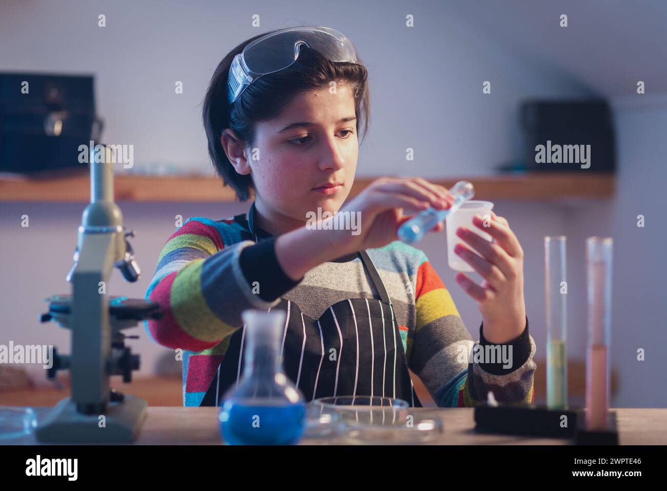 Un giovane e carino chimico in un laboratorio di ricerca domestico che indossa occhiali di sicurezza sta conducendo esperimenti con liquidi vicino a un microscopio. Avanzate Foto Stock