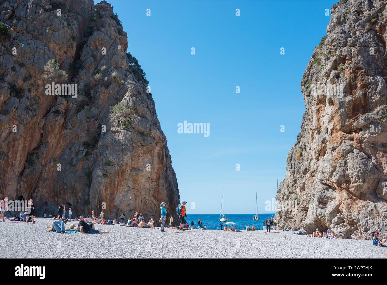 Torrent de Pareis, gente che si rilassa su una popolare spiaggia turistica di ciottoli incorniciata da alte scogliere, vista, scogliere, mare blu calmo sotto un blu nuvoloso Foto Stock