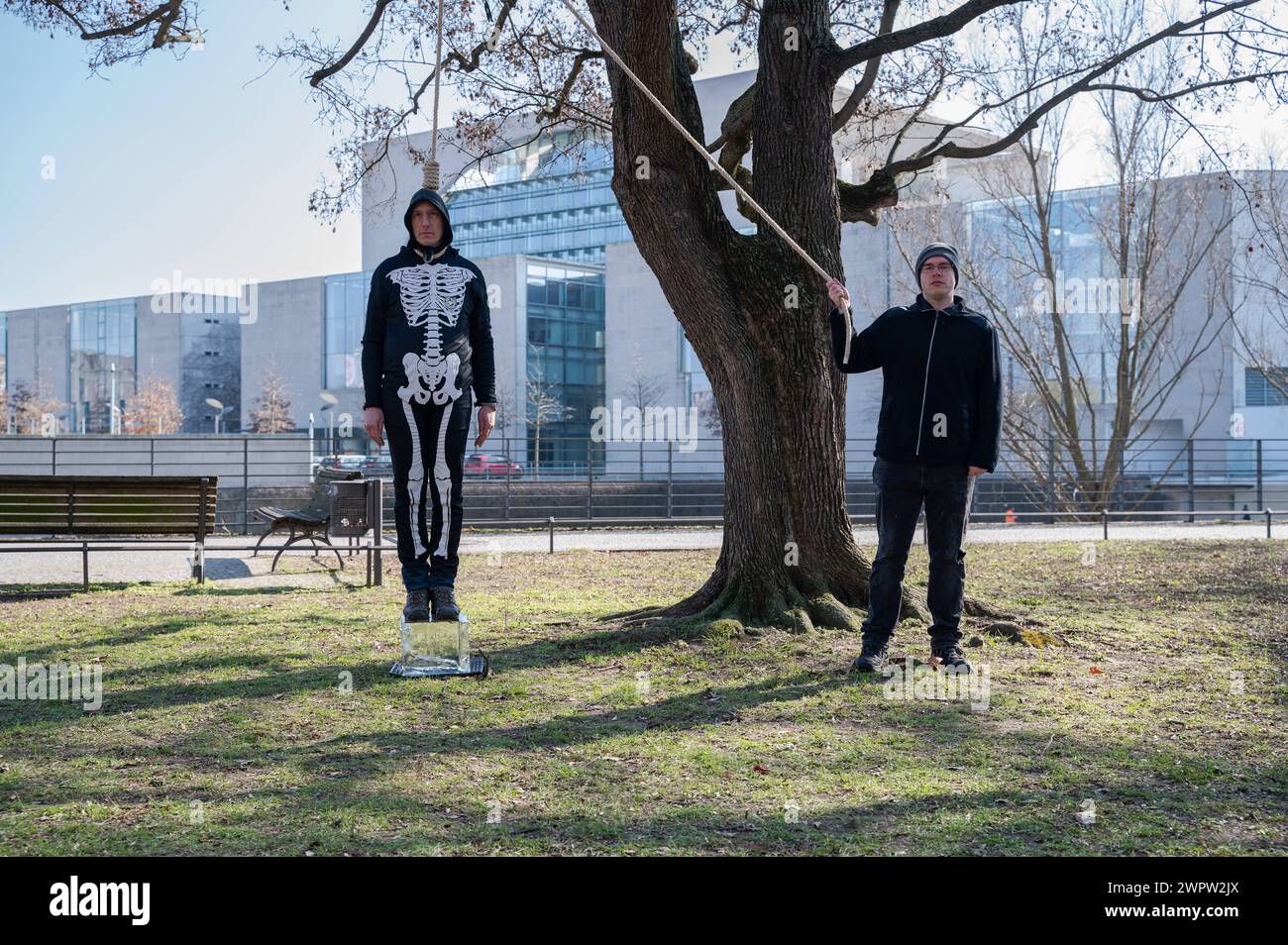 07.03.2024, Berlino, Deutschland, Europa - Der deutsche Klimaaktivist Wolfgang Wolli Metzeler-Kick traegt ein Skelettkostuem und steht mit einer Schlinge um den Hals auf einem schmelzenden Eisblock neben einem Baum, waehrend im Hintergrund das Bundeskanzleramt zu sehen ist. Der Eisblock steht symbolisch fuer die zunehmende Erderwaermung und die schmelzenden Gletscher. Schreitet die Klimaerwaermung weiterhin im jetzigen tempo voran und schmelzen die Gletscher, dann zieht sich die Schlinge um den Hals der Menschheit immer mehr zu. Der Klimaaktivist ab dem 7. Maerz 2024 in den Hungerstreik tr Foto Stock