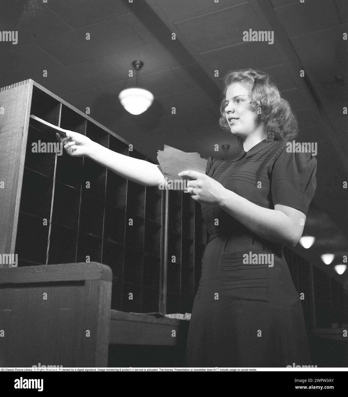 Una donna in un ufficio nel 1942. Una giovane dipendente si trova con un fascio di avvisi in mano e li ordina in diversi scomparti su uno scaffale di fronte a lei. 1942 Kristoffersson rif. B135-1 Foto Stock