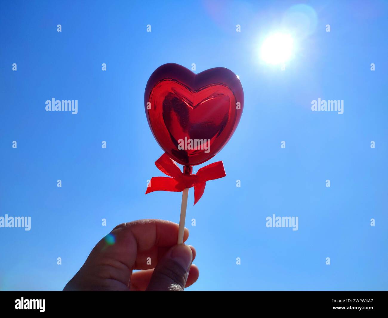 Un bastoncino a forma di cuore rosso volumetrico nella mano della donna sullo sfondo del cielo blu e del sole splendente nelle soleggiate giornate estive. Concetto di amore, San Valentino, innamorarsi, innamorati. Verticale Foto Stock