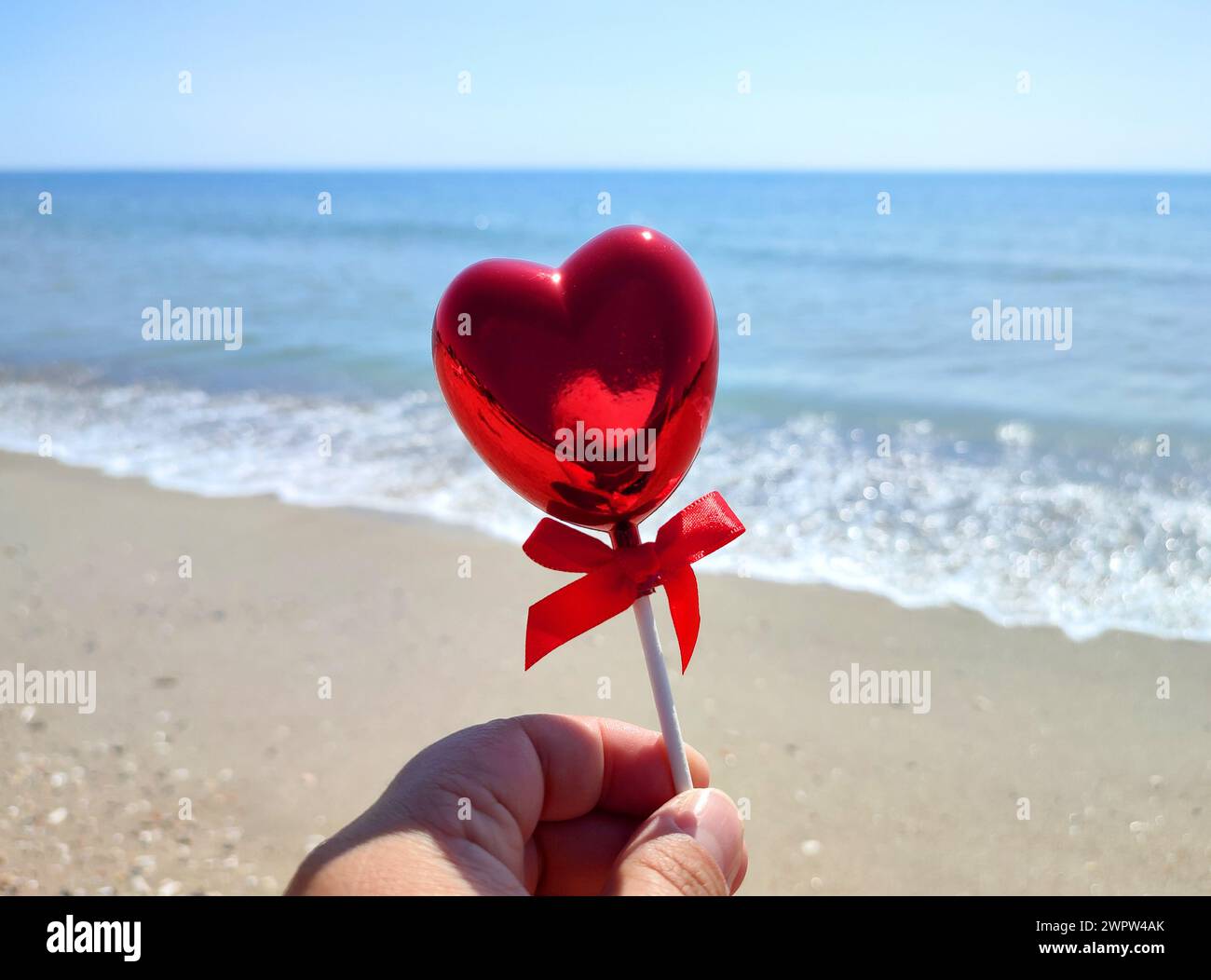 Un bastoncino a forma di cuore rosso volumetrico in mano a una donna sullo sfondo del mare blu, della spiaggia sabbiosa e del cielo blu nelle soleggiate giornate estive. Concetto di amore, San Valentino, innamorarsi. Verticale Foto Stock