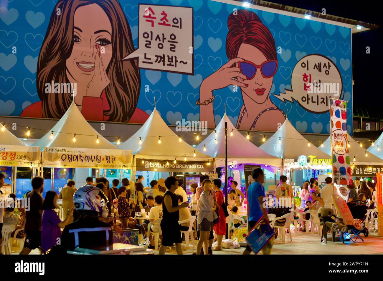 Sokcho City, Corea del Sud - 30 luglio 2019: Un mercato notturno attivo sulla spiaggia di Sokcho, sotto un grande edificio con murales e tende pop-up bianche illuminate Foto Stock