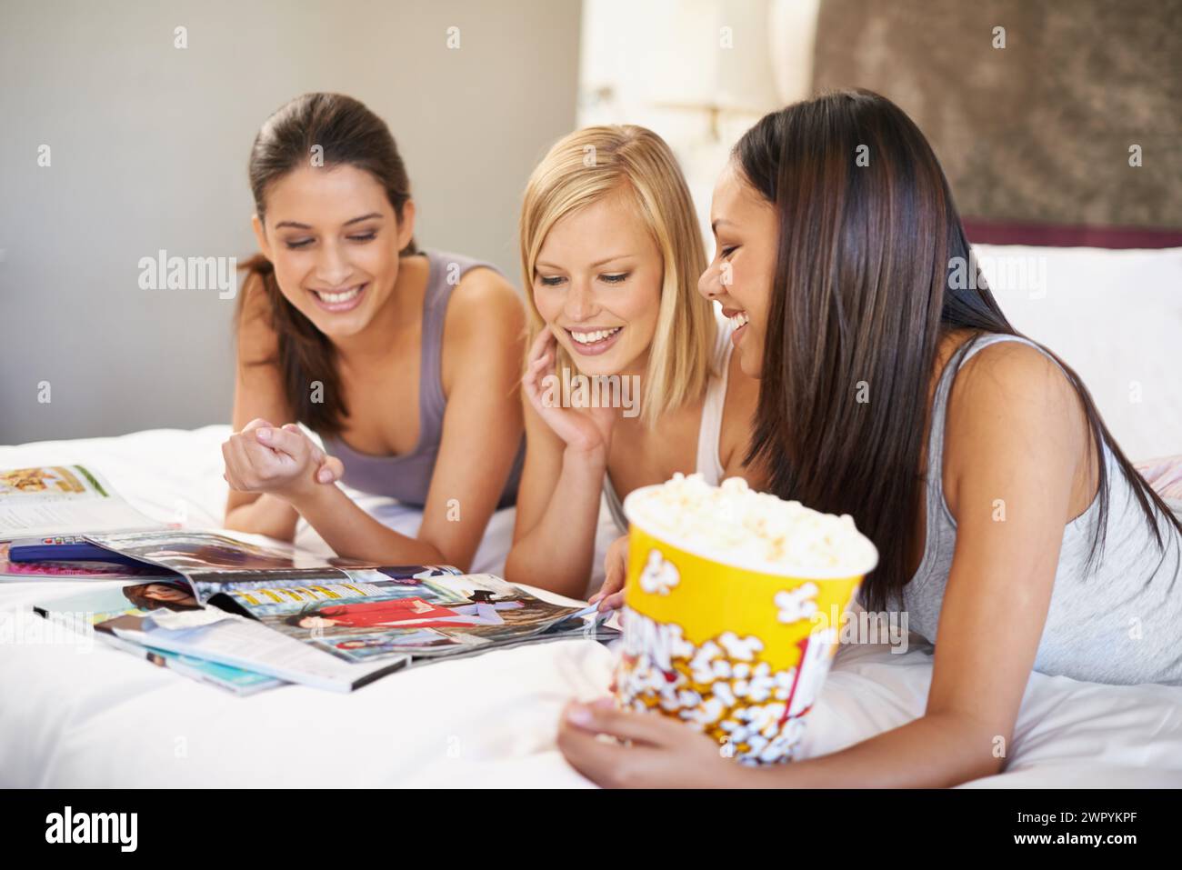 Amici, popcorn e lettura per dormire, socializzare e fare una camera da letto per legare e fare uno spuntino in gruppo. Donne, insieme e riviste per Foto Stock