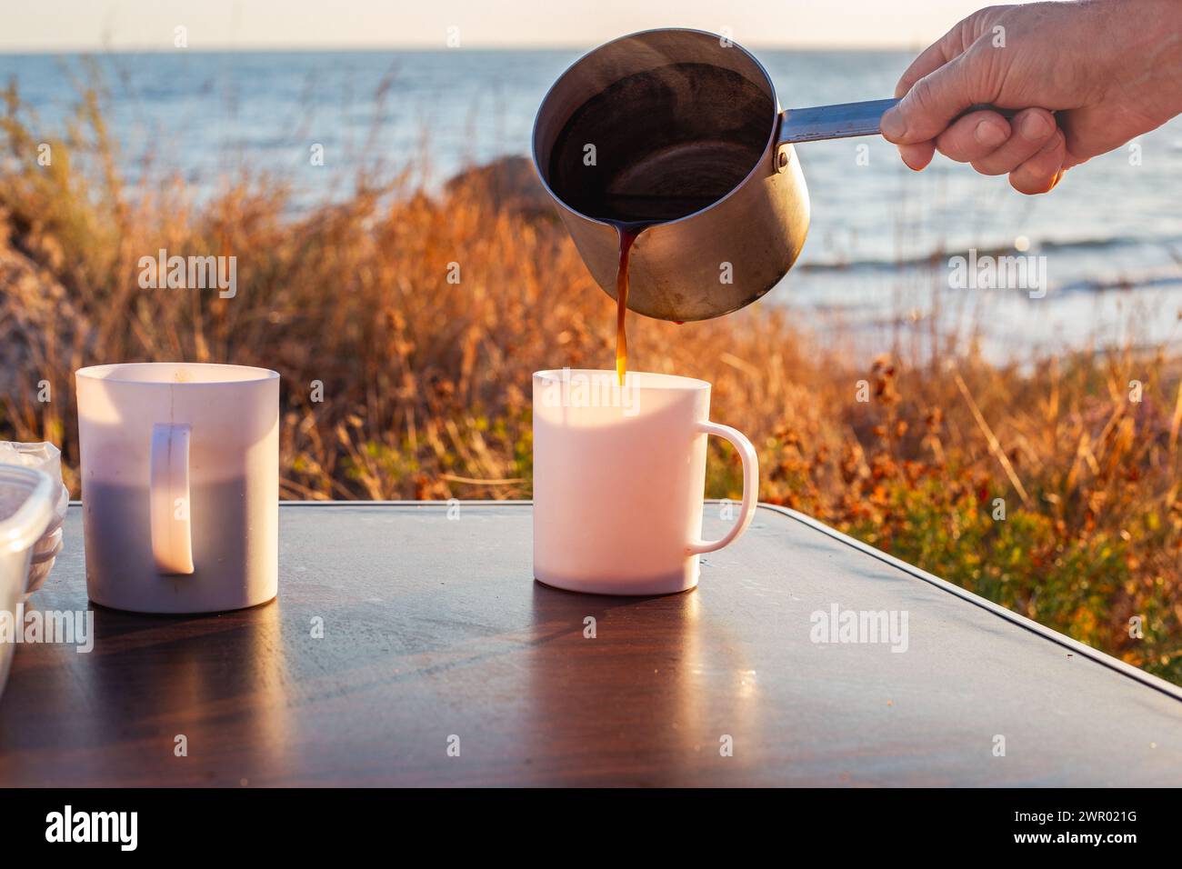 l'uomo versa il caffè appena estratto da un mestolo in alluminio in una tazza su un tavolo turistico, in riva al mare. Viaggi e turismo. Foto Stock