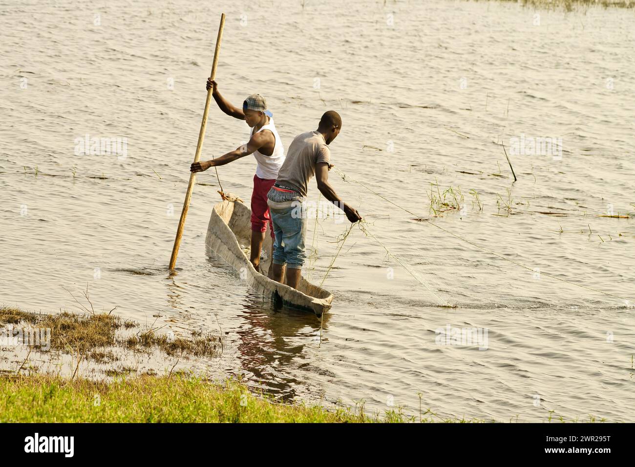 Due abitanti del villaggio che pescano da una canoa in una laguna di un fiume in Africa; uno sta colpendo la barca e uno sta lanciando la rete da pesca Foto Stock