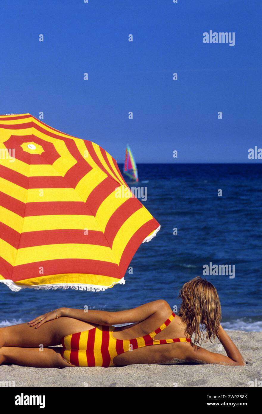 capelli biondi bella giovane donna prendere il sole sulla spiaggia sdraiarsi silhouette fotocamera posteriore davanti colorata ombrello rosso e giallo sfondo blu cielo Foto Stock