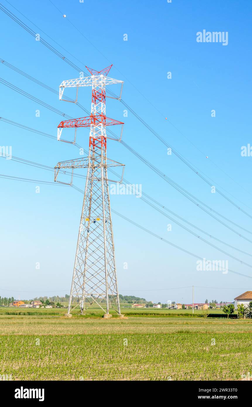 Alto pilone elettrico in un campo coltivato in una limpida giornata primaverile. Gli edifici agricoli e le case sono visibili sullo sfondo. Foto Stock