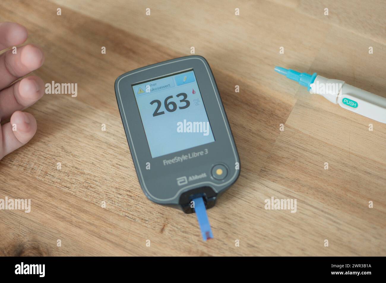 Glucometro con strisce reattive e una goccia di sangue si trova al centro di un tavolo di legno, valore elevato, sulla sinistra delle dita di un bambino, sul Foto Stock