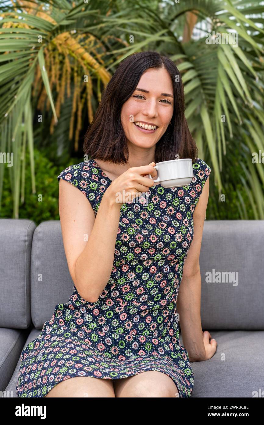 Una donna è seduta su un divano e tiene in mano una tazza di caffè. Sta sorridendo e si sta godendo il suo drink ahile guardando la macchina fotografica Foto Stock