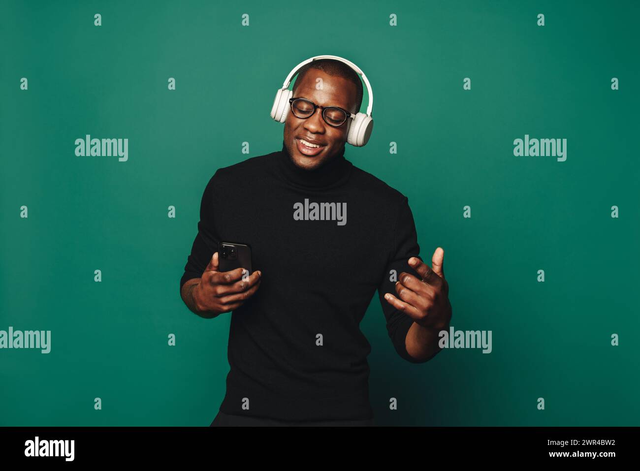 Il giovane con occhiali e abbigliamento casual balla con gioia su sfondo verde. Ha in mano uno smartphone e indossa un auricolare elegante per ascoltare Foto Stock