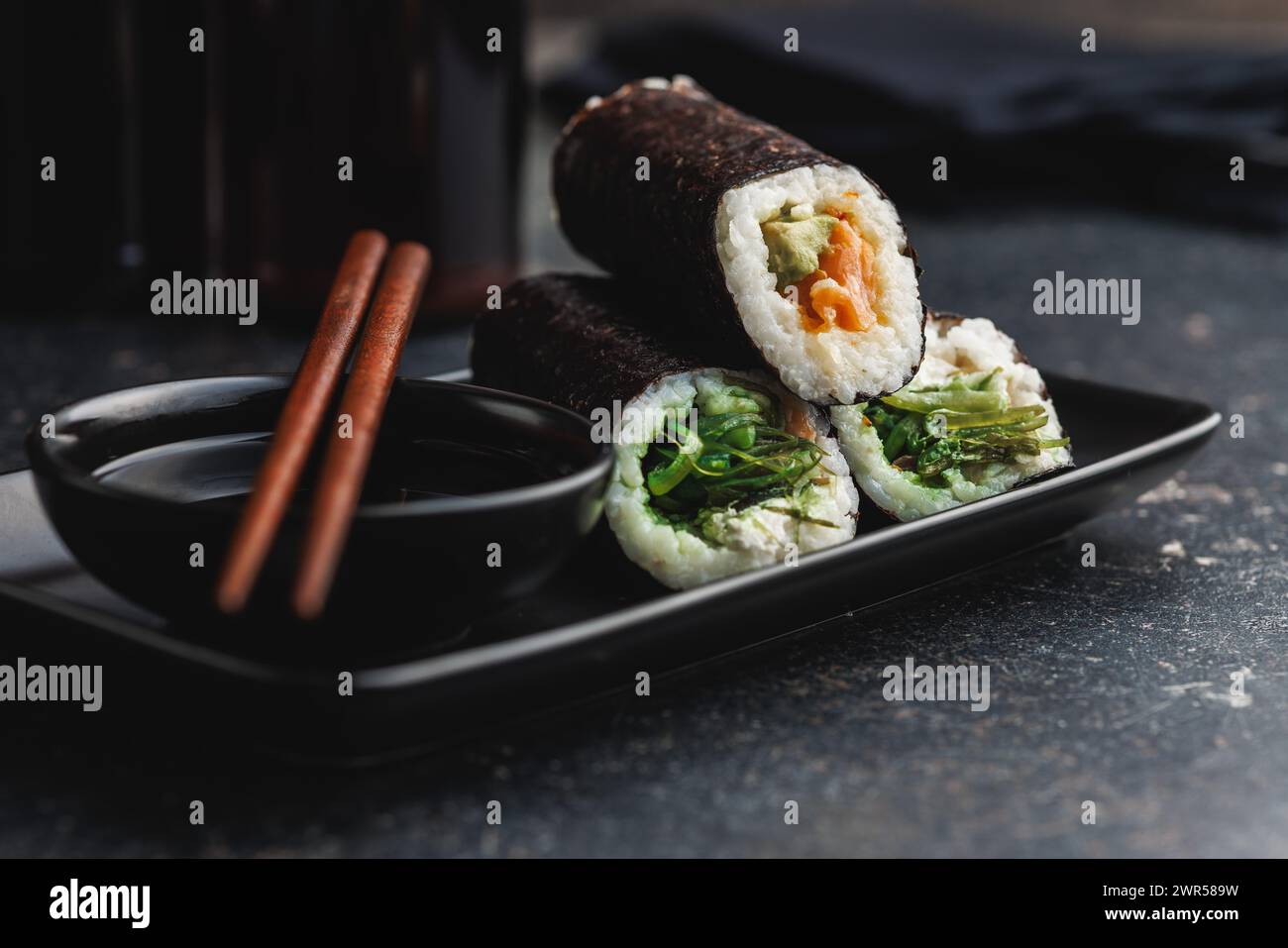 Un piatto nero con pezzi di sushi con bacchette e ciotola di salsa di soia. Il sushi mostra una varietà di colori, texture e ripieni, in vetrina Foto Stock