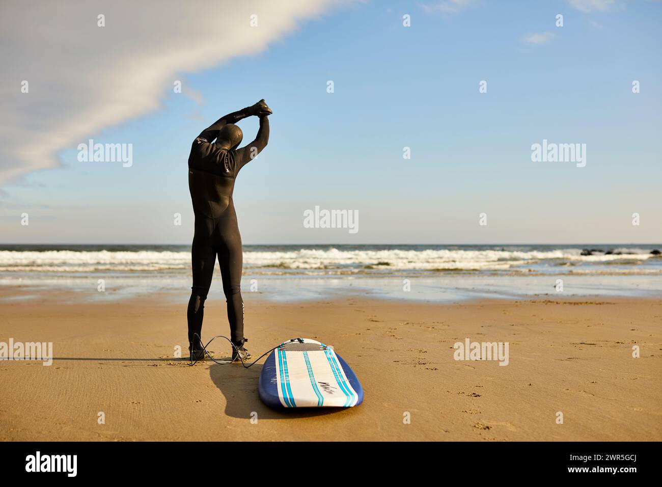 Un surfista maschile che si allunga e si riscalda prima di fare surf in spiaggia Foto Stock