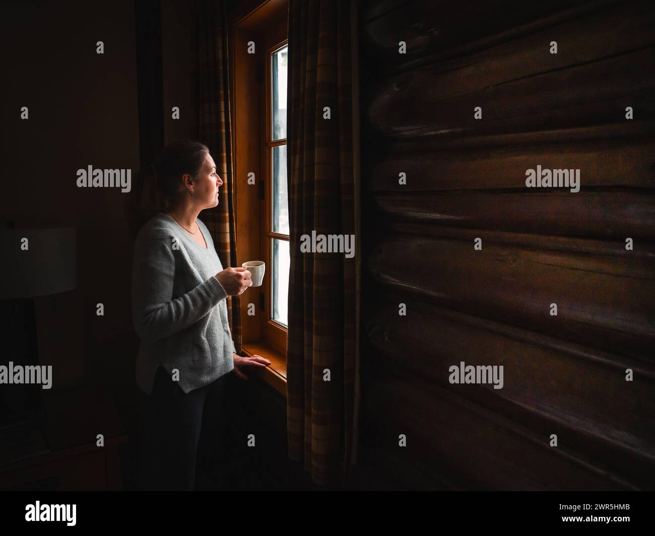 Donna che tiene una tazza di caffè guardando fuori dalla finestra della cabina di legno. Foto Stock