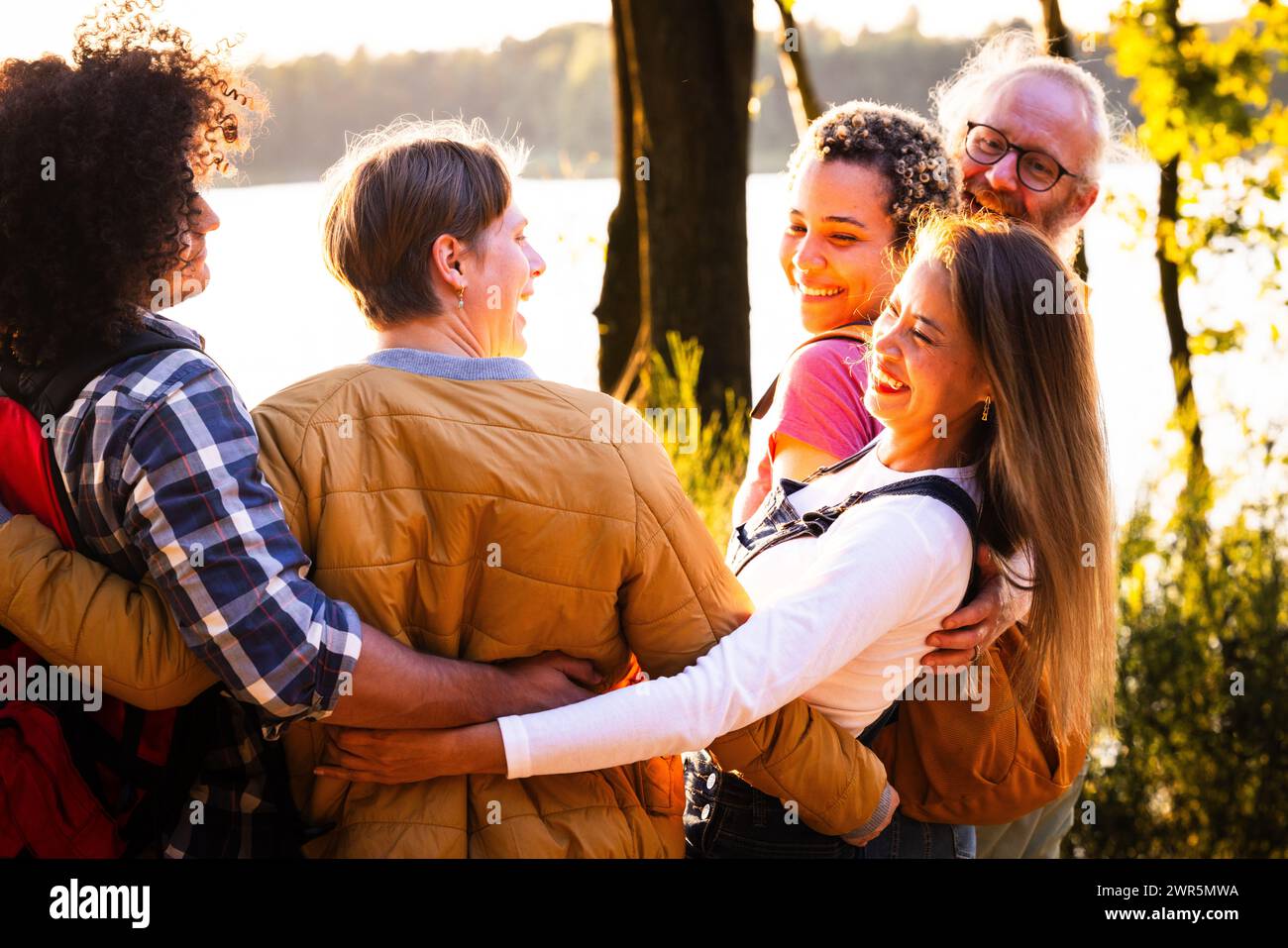 Questa immagine trasuda calore e cameratismo mentre un gruppo di amici multietnici condivide un abbraccio di gruppo sul lago durante il tramonto. I loro volti sorridenti e la vicinanza del loro abbraccio riflettono forti legami e felicità. La retroilluminazione del sole che tramonta crea un effetto alone intorno a loro, aggiungendo un senso di gioia e pace al raduno. Il variegato gruppo, compresi uomini e donne di età e background etnici diversi, illustra l'inclusività e la bellezza dell'amicizia. Gruppo abbracciato tra amici multietnici vicino al lago Suns. Foto di alta qualità Foto Stock