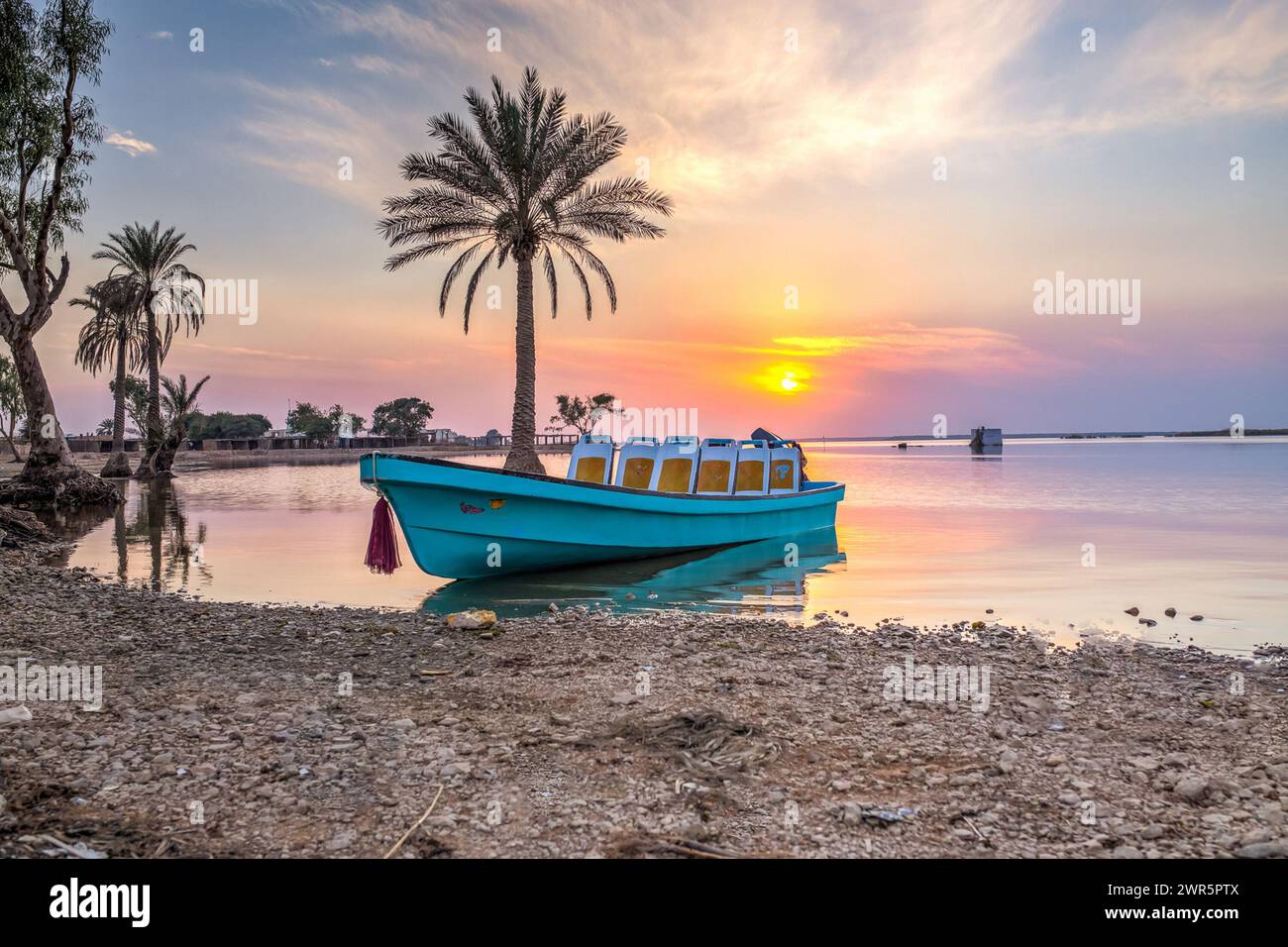 Una barca blu solitaria galleggia vicino al riflesso della palma mentre il sole tramonta, gettando calde sfumature sulle acque calme. Foto Stock