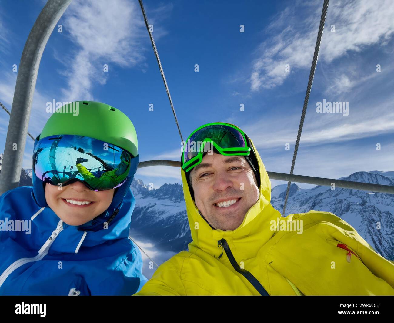 Un paio di sciatori felici adulti e un bambino, indossando giacche da sci luminose, occhiali di protezione, catturano un momento allegro su uno skilift con le bianche vette alle spalle Foto Stock