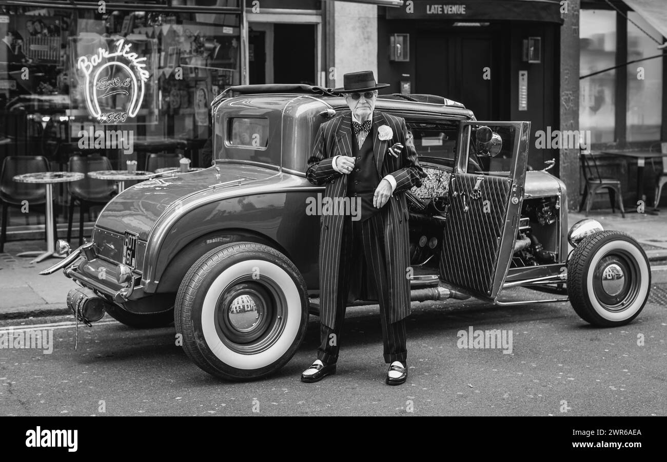 Un'immagine in bianco e nero di un uomo ben vestito in piedi accanto a un'auto d'epoca fuori Bar Italia a Londra, a Soho. Foto Stock