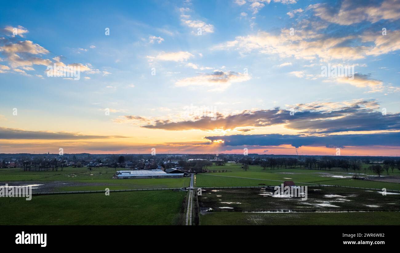 Mentre il sole tramonta, i suoi raggi sbirciano attraverso la copertura delle nuvole, gettando una luce spettacolare su vasti terreni agricoli e strutture sottostanti. Cielo al tramonto sui terreni agricoli: Una maestosa esposizione di nuvole e luci. Foto di alta qualità Foto Stock