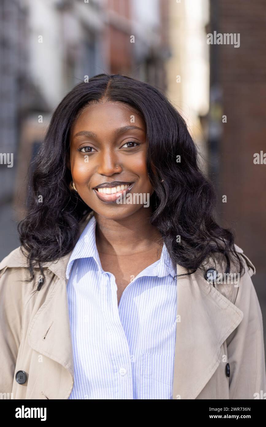 L'immagine è un ritratto ravvicinato di una giovane donna africana con un sorriso radioso, posizionato su uno sfondo urbano. I suoi capelli cadono in onde morbide intorno alle sue spalle ed è elegantemente vestita con una camicia a righe blu sotto un classico trench beige. Il suo look è sia professionale che accessibile, perfetto per l'ambiente urbano in cui si trova. Lo sfondo è leggermente sfocato, attirando tutta l'attenzione sulla sua espressione coinvolgente. Sorriso radioso di una giovane donna africana in città. Foto di alta qualità Foto Stock