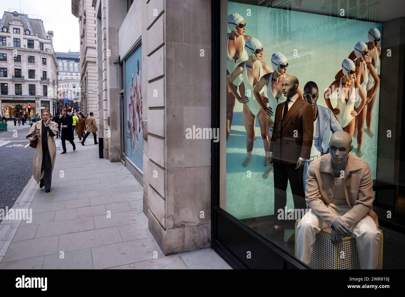 Immagini pubblicitarie con diverse donne in costume da bagno in formazione per il rivenditore di moda Suit Supply che interagiscono con manichini maschili nella vetrina del negozio mentre le persone passano a Mayfair il 6 marzo 2024 a Londra, Regno Unito. Foto Stock