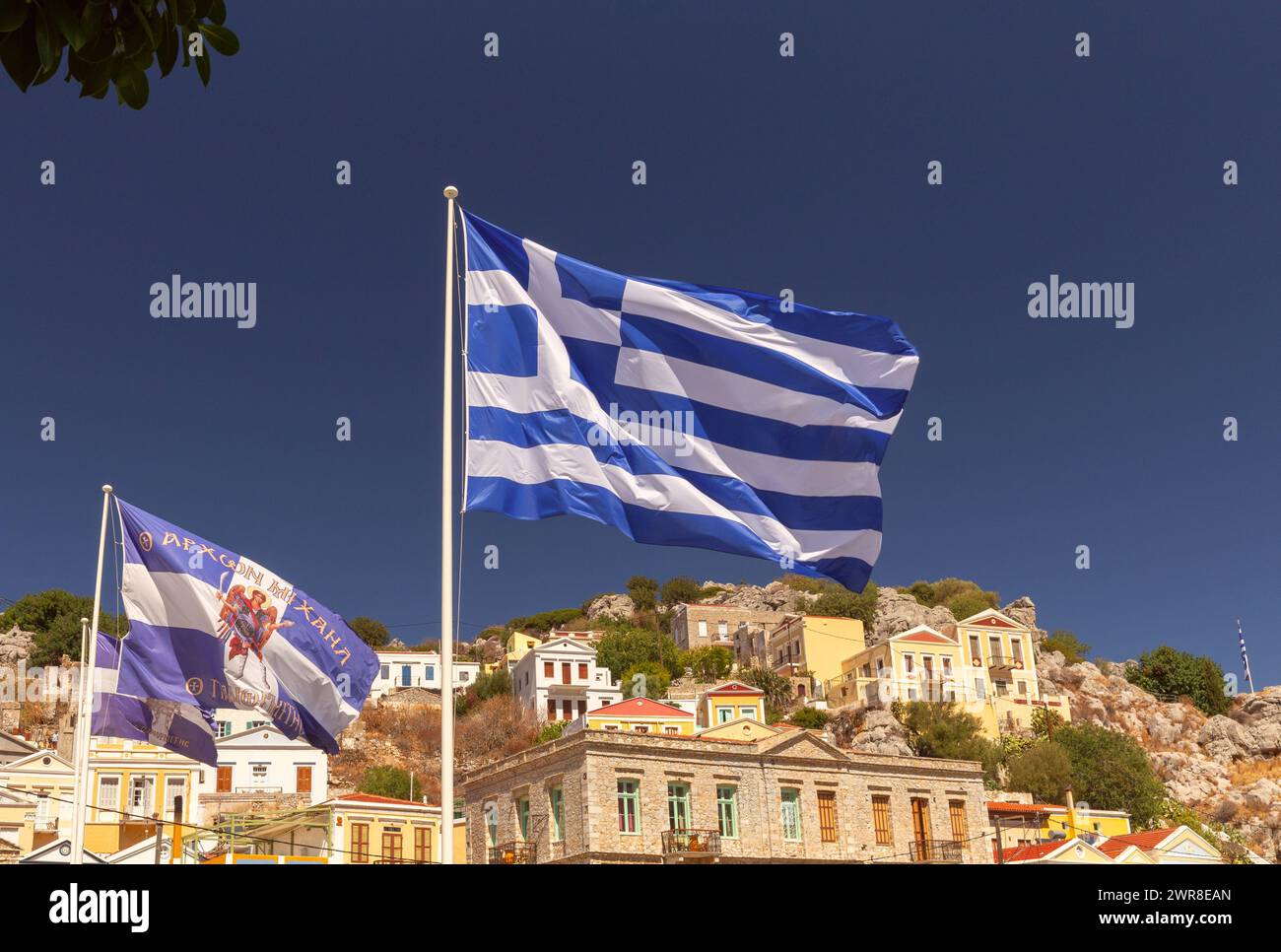 La bandiera nazionale greca sullo sfondo del cielo blu delle montagne e le facciate delle vecchie case nel villaggio di Symi. Grecia. Foto Stock