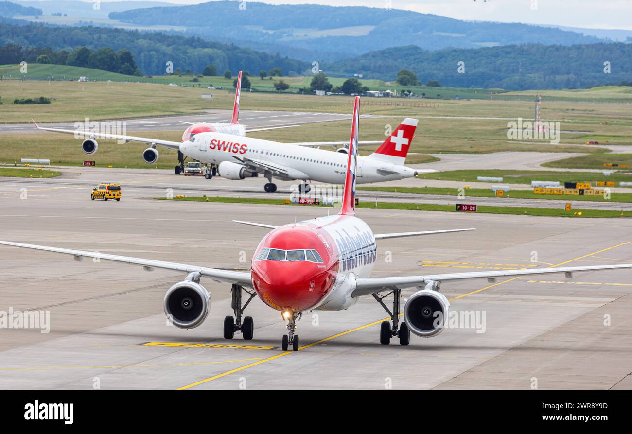 Vorne rollt ein Airbus A320-214 nach der Landung zum Terminal auf dem Flughafen Zürich. Dahinter wird ein vierstrahliger Airbus A340 von einem Schlepp Foto Stock