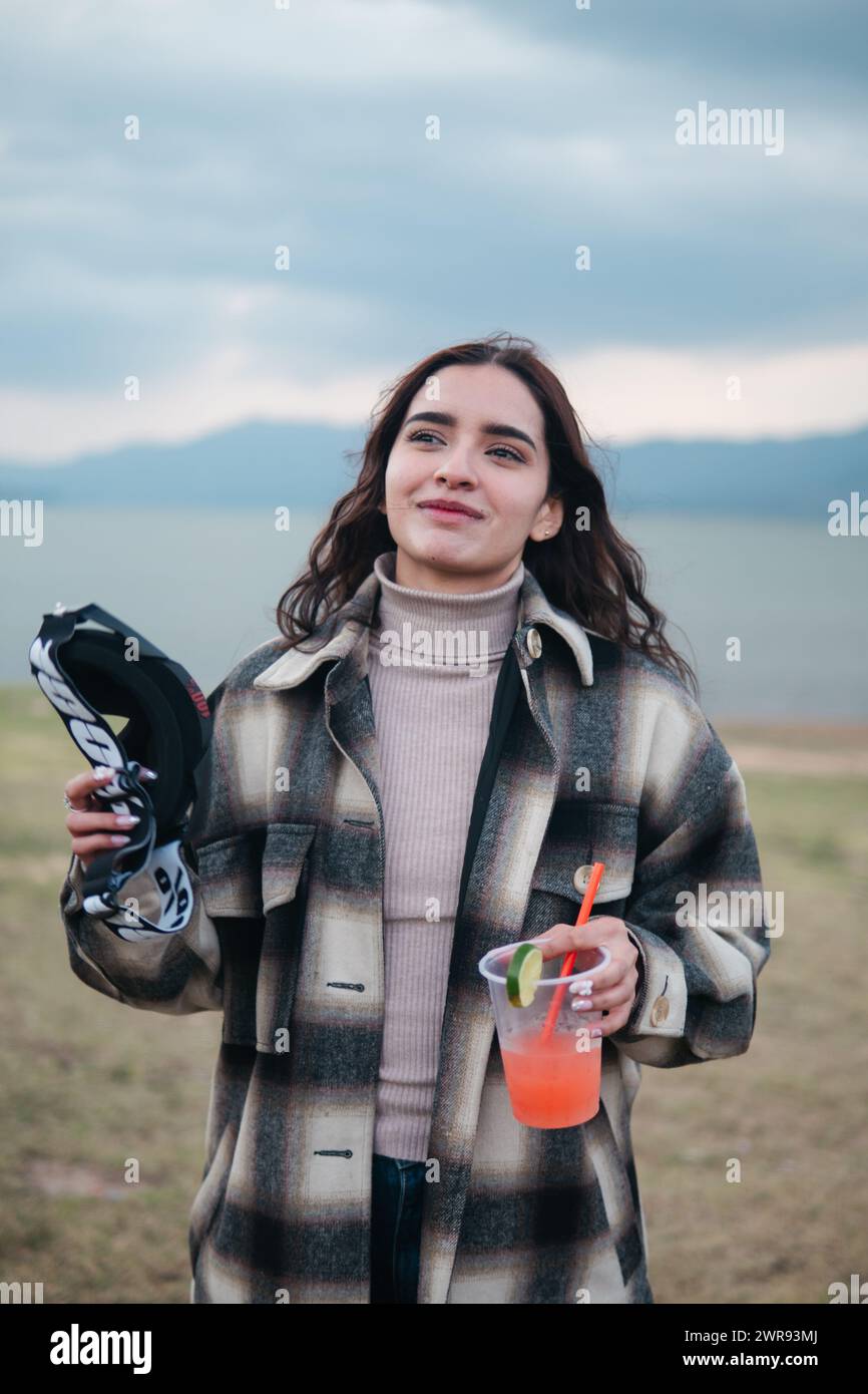 Una giovane donna sorridente regge occhiali da motocross e un cocktail fresco, in piedi in un campo aperto con un lago sullo sfondo Foto Stock