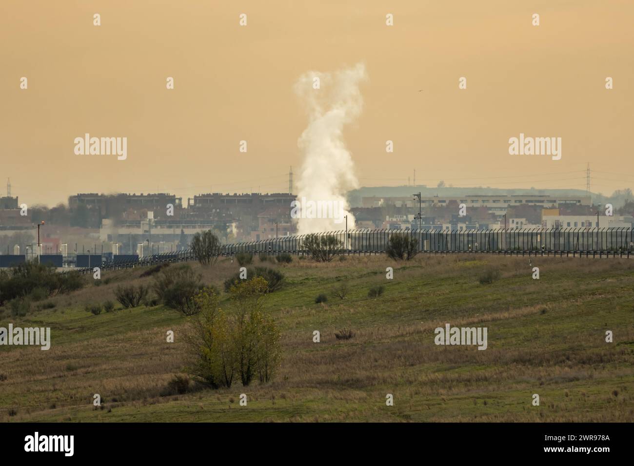 Una colonna di vapore al centro del campo accanto a una recinzione con la vista di una città sullo sfondo Foto Stock