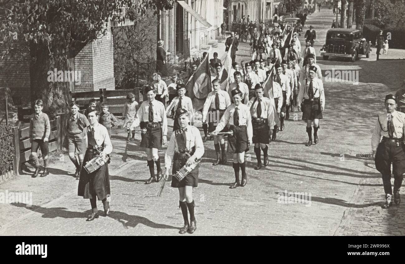 Youth Storm sfilano con la batteria, Youth Storm (titolo della serie), gruppo di membri della Youth Storm, con una ragazza e un ragazzo con un tamburo davanti, cammina attraverso una strada con ville. Dietro di loro solo ragazzi, due con una tromba, e alcuni con una bandiera, quello olandese e quello della tempesta di Jeugdstorm. Tutto in uniforme. L'uomo all'estrema destra con le breeches potrebbe essere il fotografo amatoriale Van Schaaik. Alcuni bambini piccoli camminano con loro. Dietro un'auto sulla destra., J. otto, Paesi Bassi, 1935 - 1937, supporto fotografico, stampa in gelatina argento, altezza 8,5 cm x larghezza 13,5 cm Foto Stock