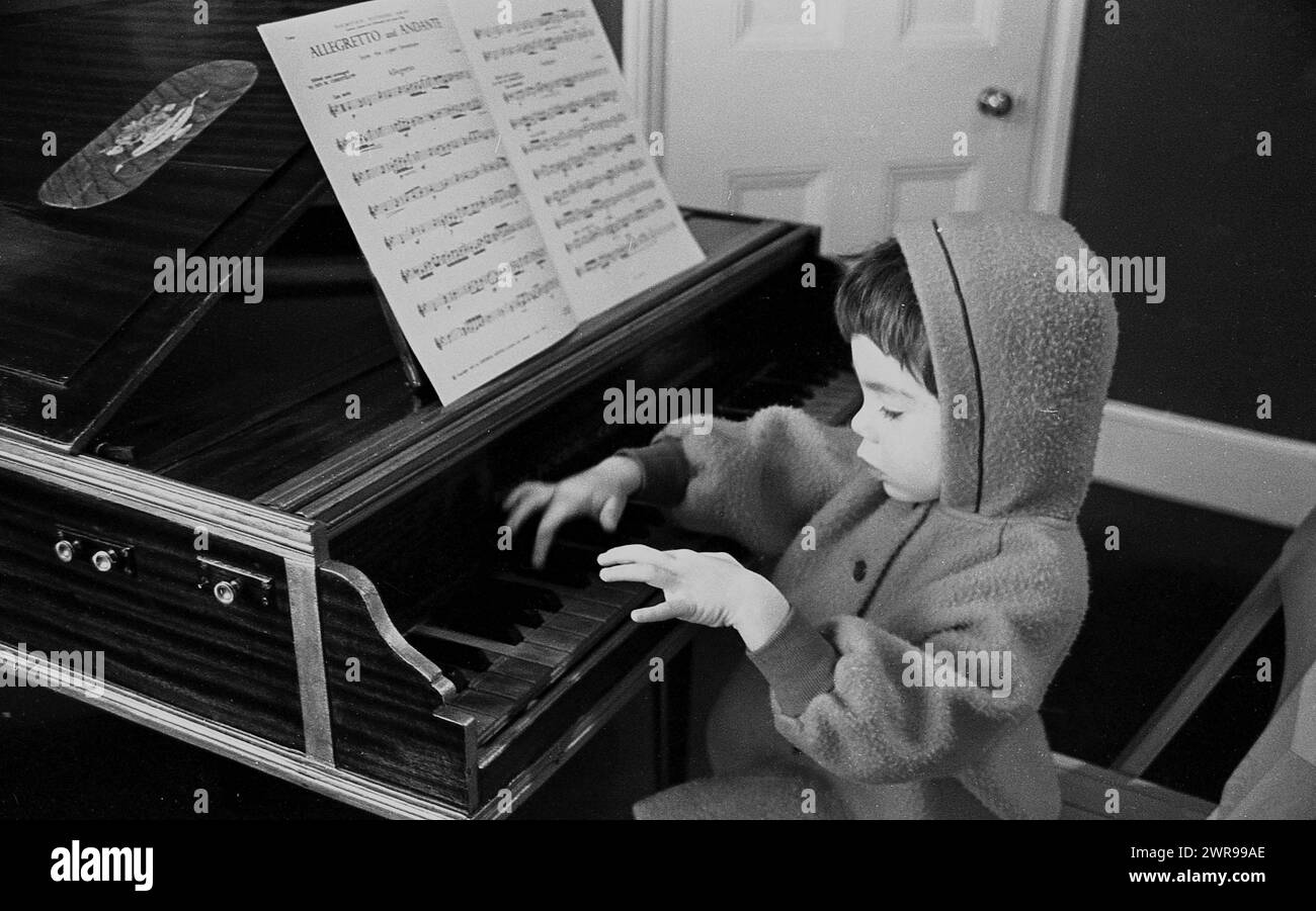 Anni '1960, storico, un ragazzino in pile con cappuccio che si staglia davanti a un pianoforte per bambini premendo un tasto, spartiti davanti a lui, Inghilterra, Regno Unito. Foto Stock