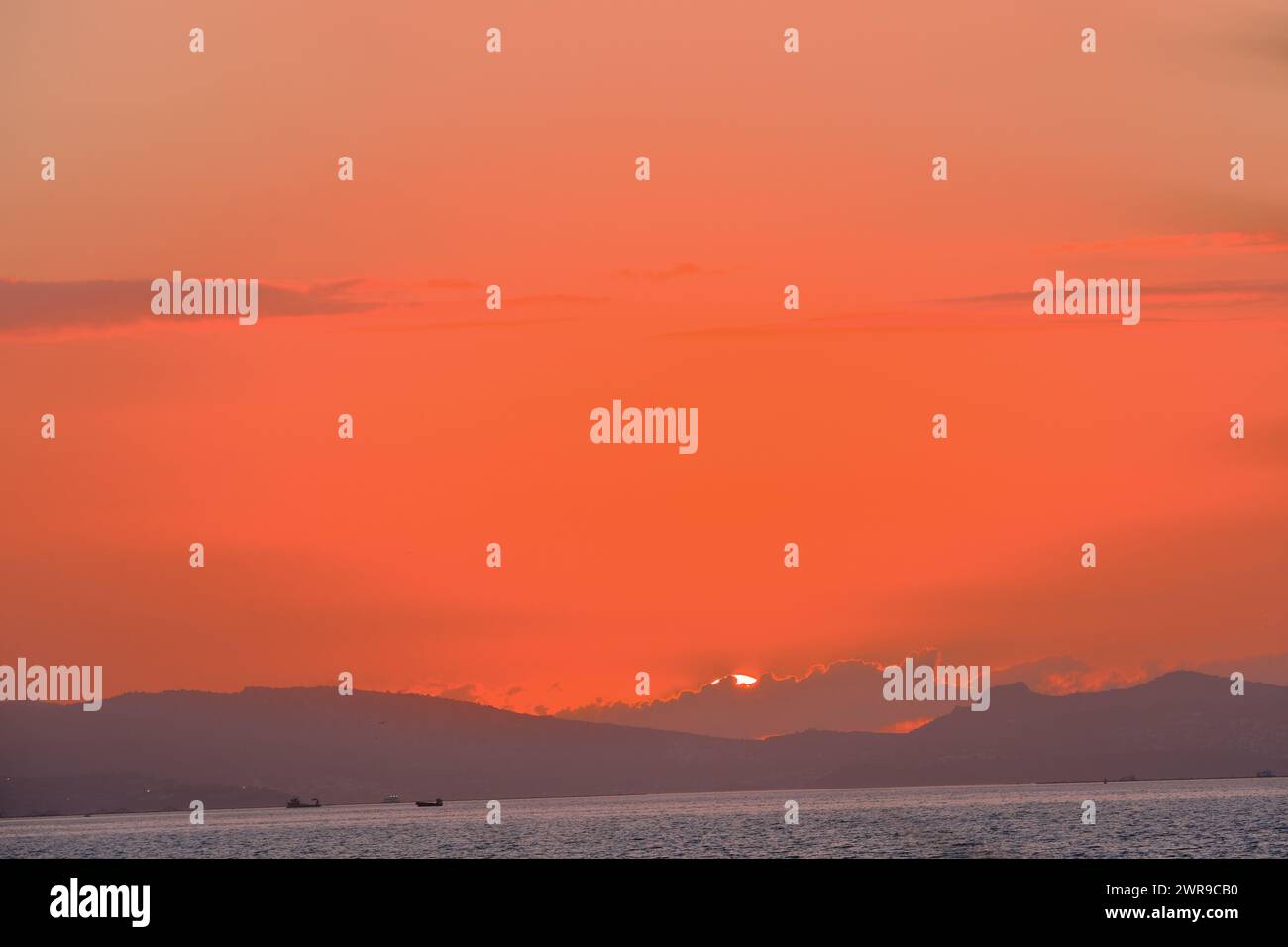 Cielo cremisi e sole a sfera al tramonto sul mare dietro le colline Foto Stock
