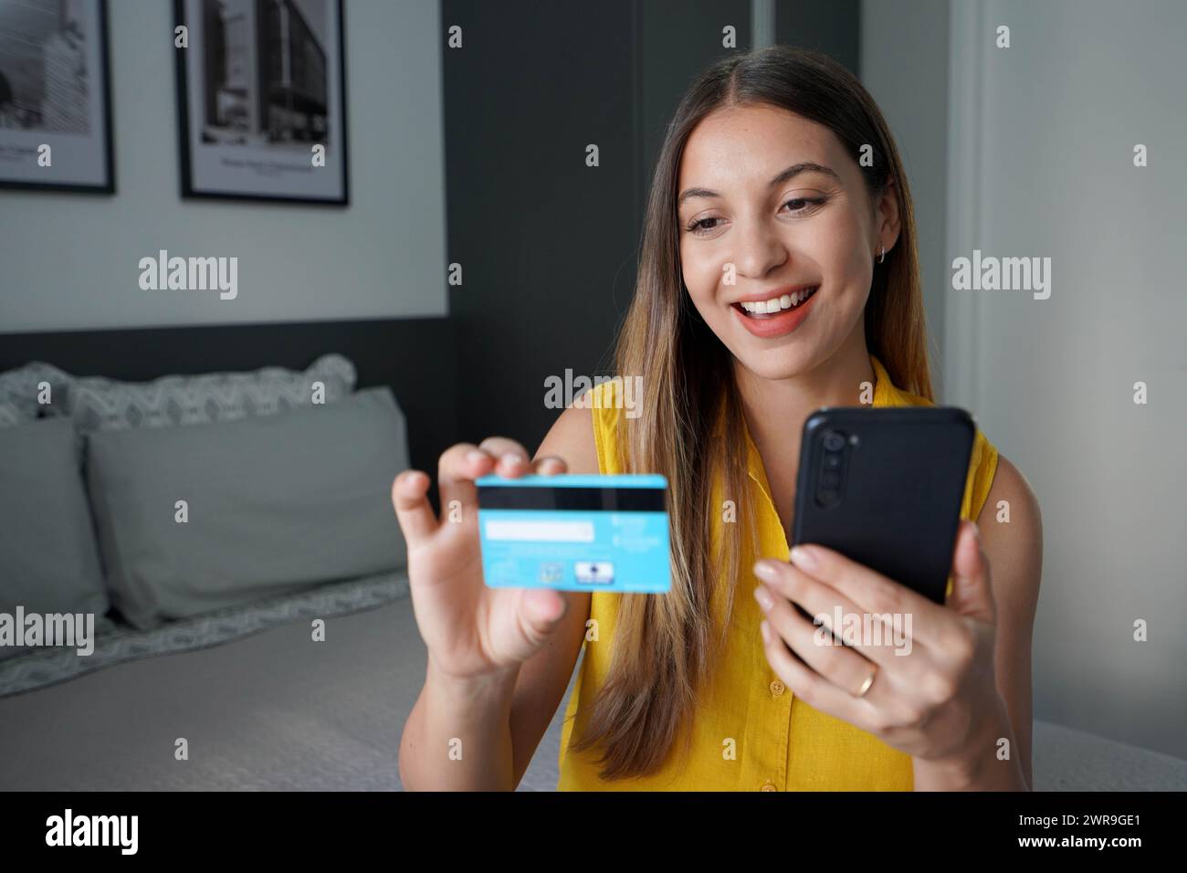 Ritratto di una ragazza che ride leggendo il numero di carta di credito per il pagamento online e tenendo il suo smartphone in camera da letto a casa Foto Stock