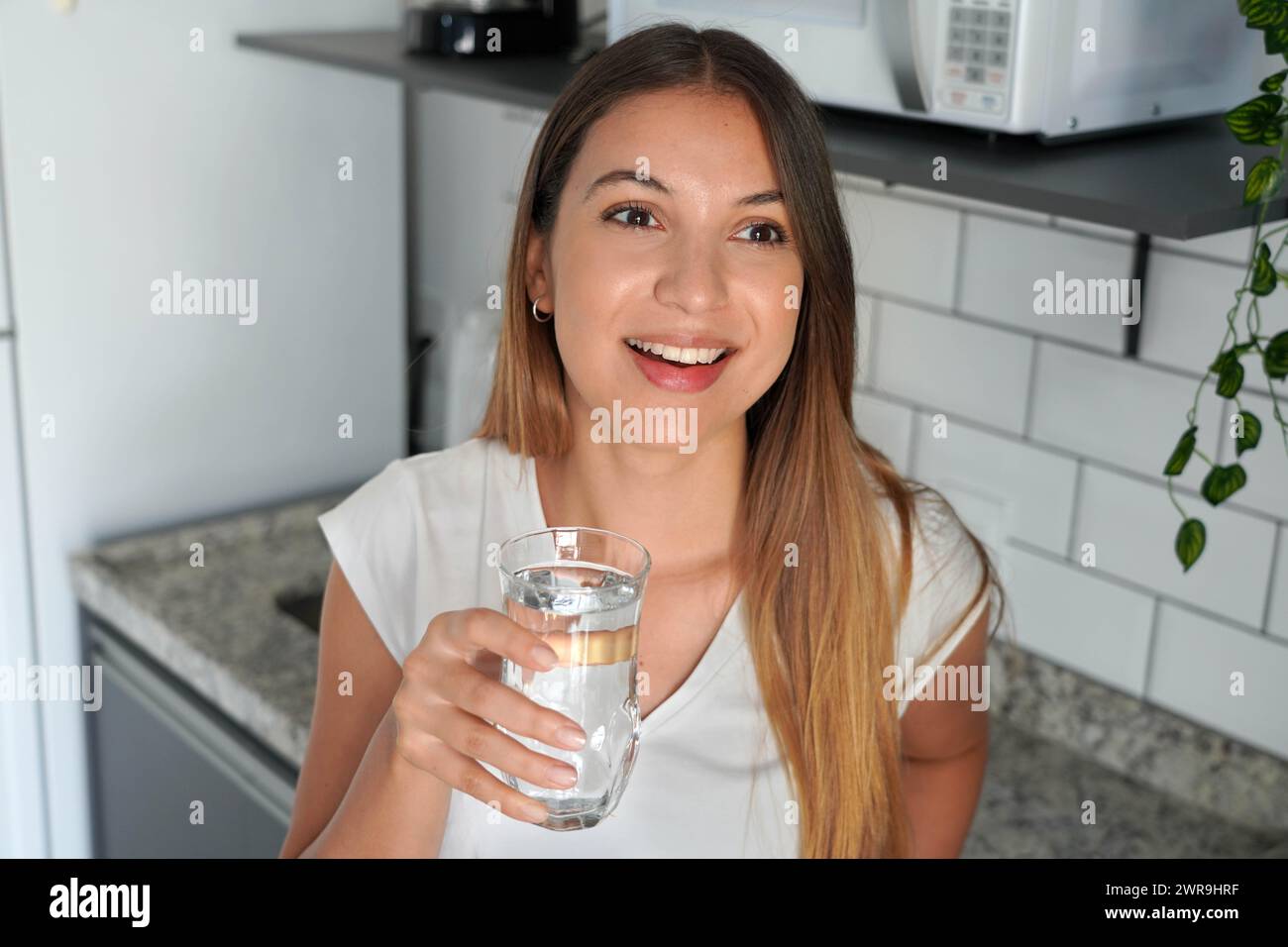 Bella giovane donna che tiene un bicchiere pieno d'acqua in cucina a casa Foto Stock