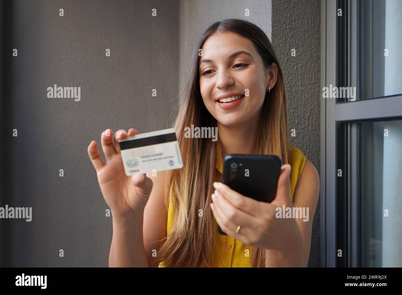 Felice giovane donna brasiliana che tiene in mano lo smartphone e legge il numero di carta di credito per lo shopping online Foto Stock