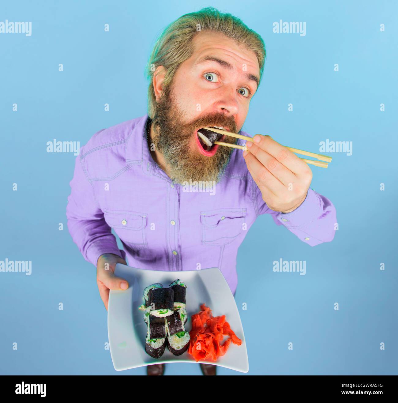 Uomo barbuto che mangia un gustoso sushi roll con bacchette. Menu di sushi. Bell'uomo in camicia in denim con piatto delizioso maki sushi e zenzero sottaceto Foto Stock