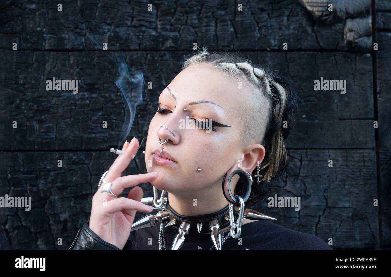 giovane ragazza gotica sexy e seducente con piercing, indossa una t-shirt teschio e un grande soffocatore di picchi, fuma una sigaretta arrotolata a mano davanti a una bruciata, c Foto Stock
