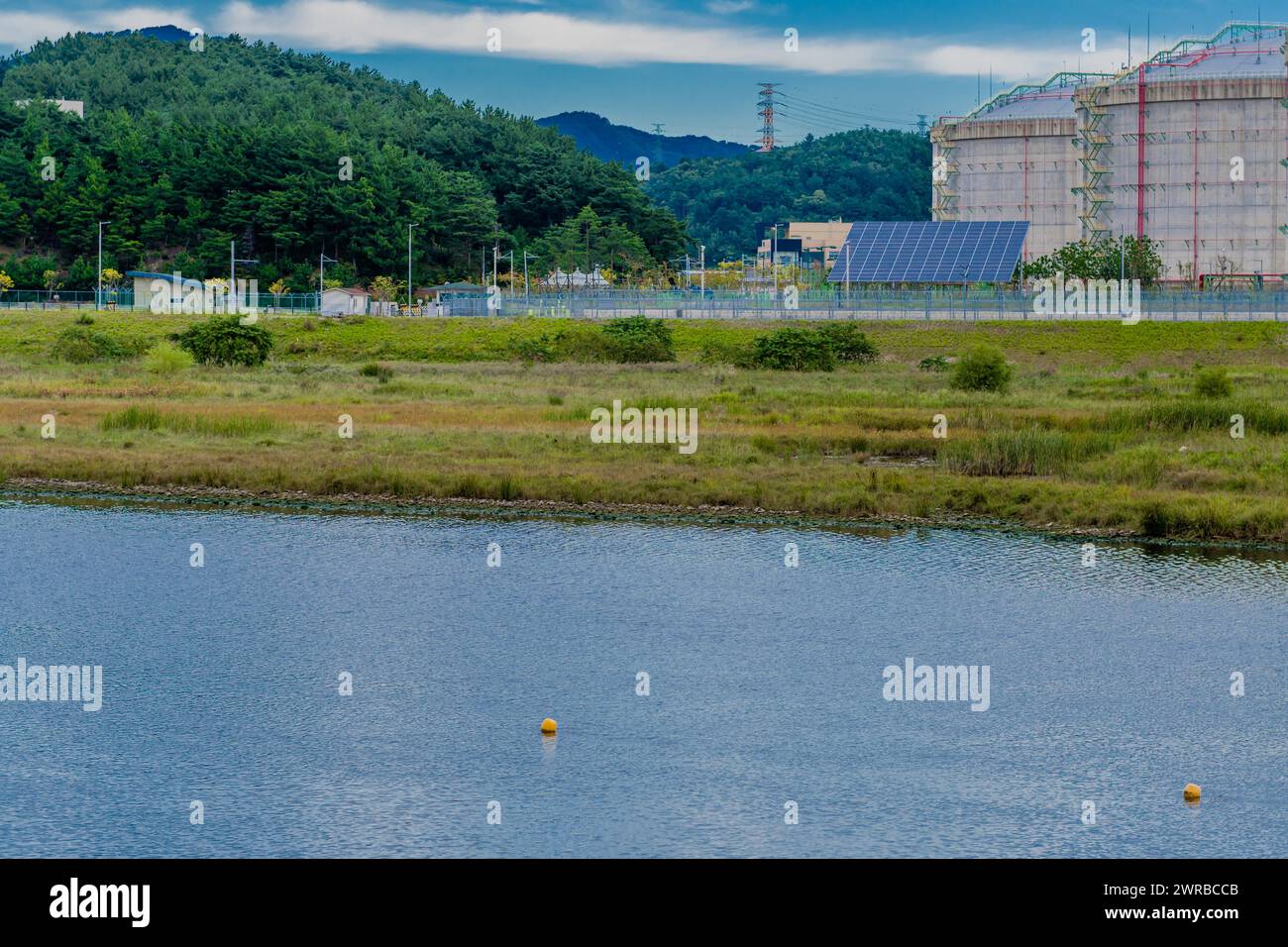 Serbatoi di stoccaggio industriali accanto a un corpo d'acqua calmo punteggiato di boe, sullo sfondo di una montagna, in Corea del Sud Foto Stock