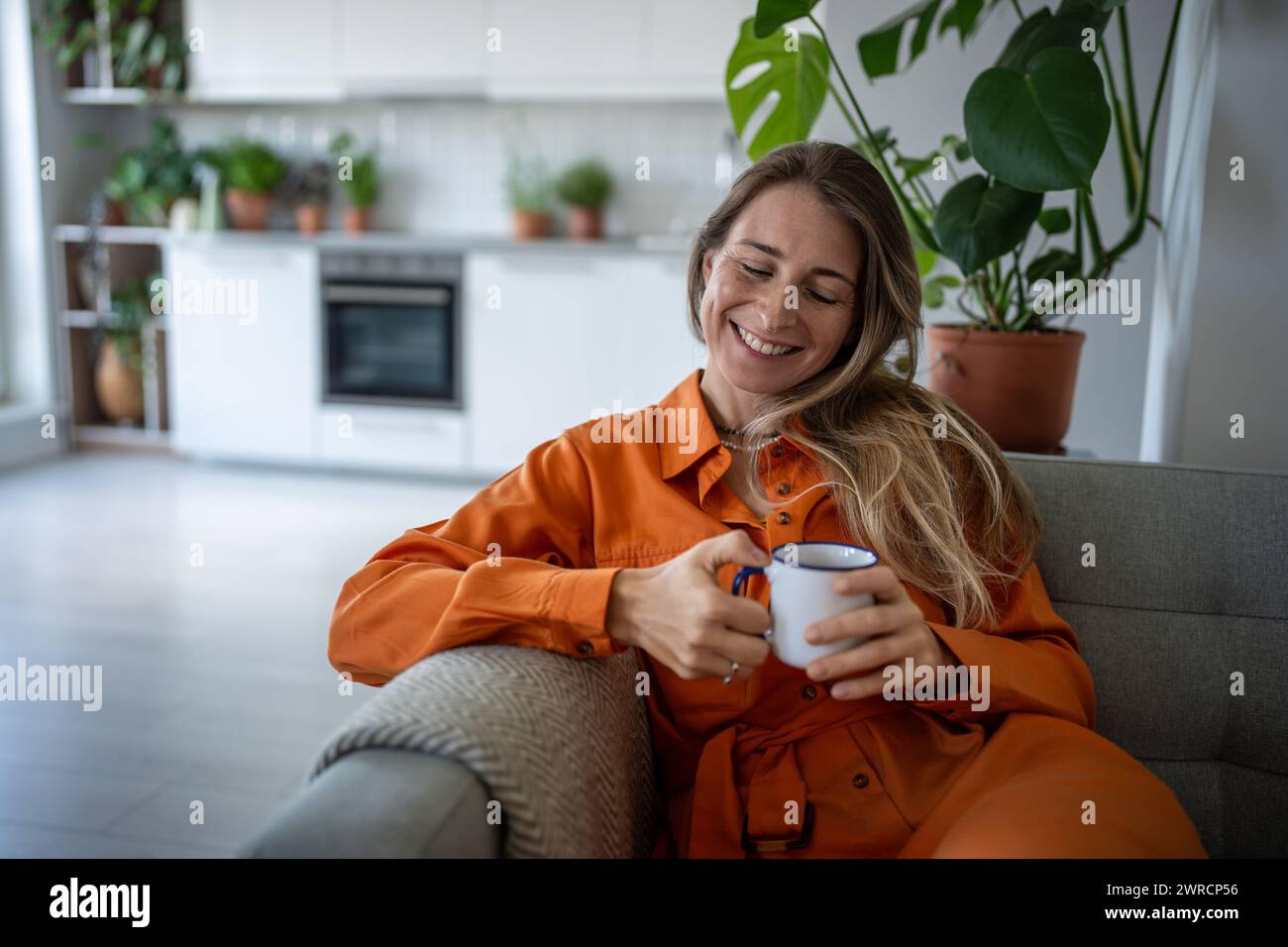 Una donna allegra con un sorriso ricorda i momenti felici del passato mentre si siede con una tazza di tè sul divano di casa. Foto Stock