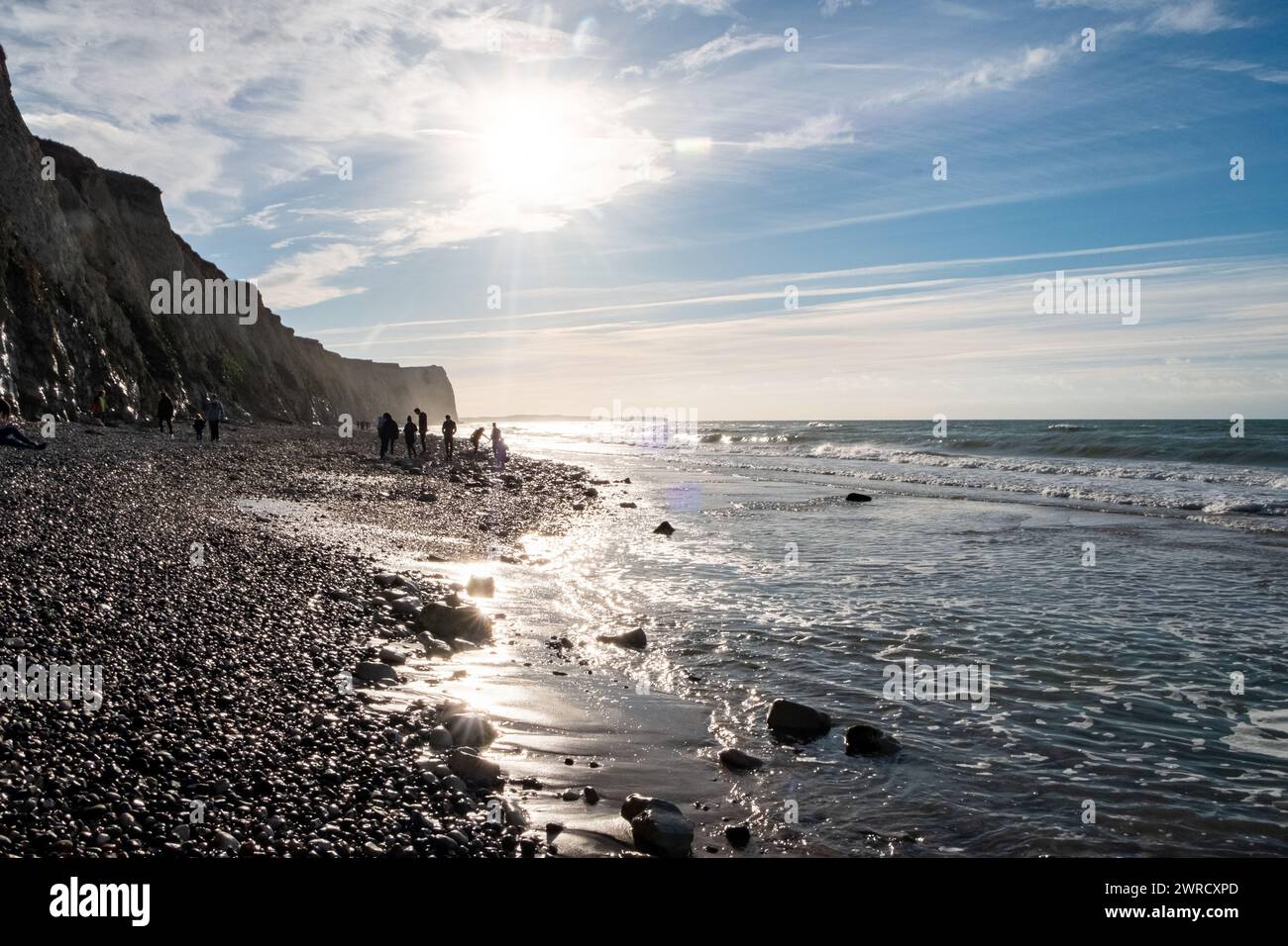 Si tratta di una suggestiva fotografia marina che cattura un tranquillo tramonto su una spiaggia di ciottoli. Il sole è posizionato in basso nel cielo, emettendo una luce soffusa che si riflette sulle pietre bagnate e sulle onde dolci, creando un'atmosfera serena. Le scogliere sulla sinistra suggeriscono una costa frastagliata, probabilmente indicativa di un luogo noto per la sua bellezza naturale. Le persone sono visibili a distanza, godendosi la spiaggia, che aggiunge un elemento umano dinamico alla scena. Servizi costieri al tramonto. Foto di alta qualità Foto Stock