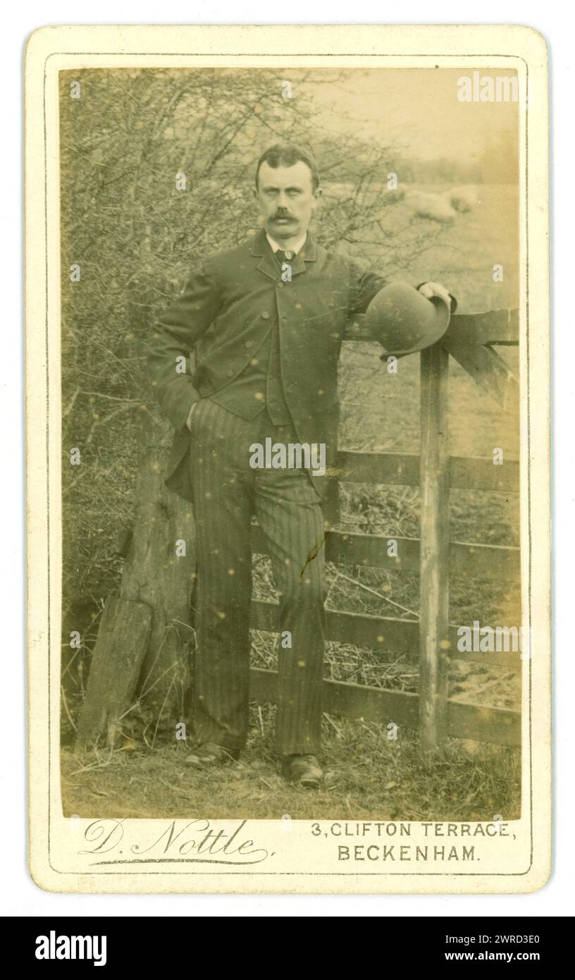 Foto originale degli anni 1880 in tono seppia - Carte de Visite vittoriana (biglietto da visita o CDV) di un uomo affascinante, forse un gentiluomo agricoltore, in abito che tiene un cappello a cilindro, fotografato in un ambiente rurale, vicino a un campo di pecore da D. Nottle del 3 Clifton Terrace, Beckenham, Kent, Regno Unito. Circa anni 1880 Foto Stock