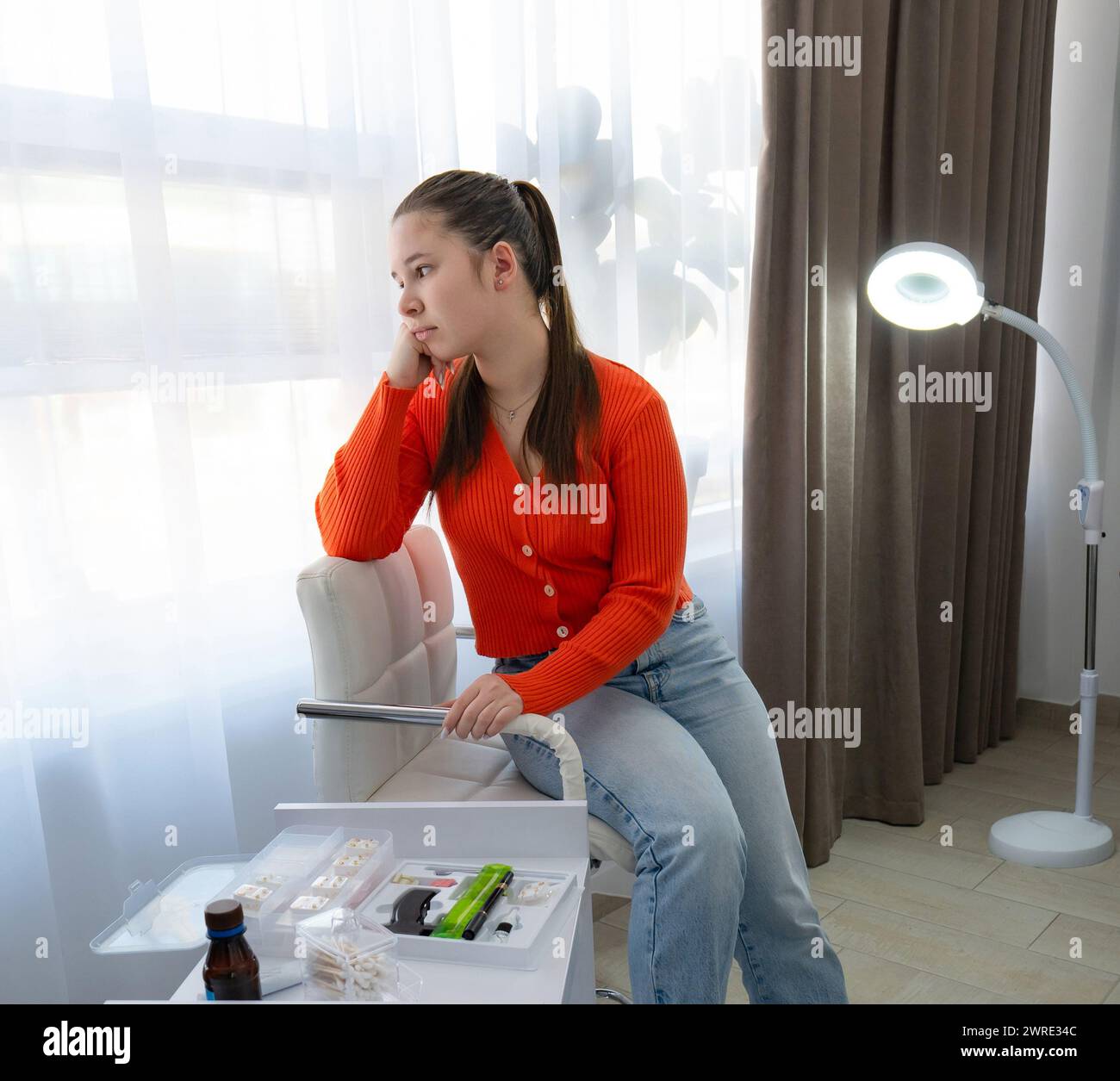 Una ragazza caucasica guarda con rabbia alla finestra, prima della procedura di piercing al salone di bellezza. Foto di alta qualità Foto Stock